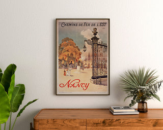 Manifesto vintage Nancy, Chemins de fer de l’Est, incorniciato sopra una credenza in legno, tra delle piante.